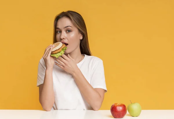 Rapariga comendo hambúrguer em vez de maçãs que jazem na mesa — Fotografia de Stock