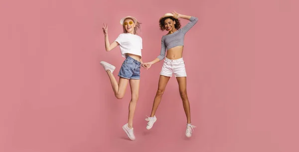 Two attractive young girls in summer clothes jumping on pink background, panorama — Stockfoto