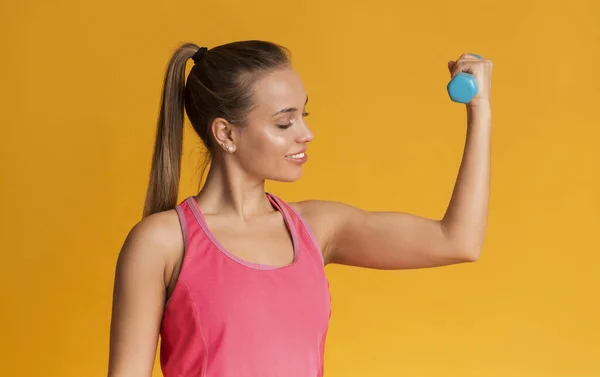 Chica joven deportiva demostrando bíceps, haciendo ejercicio con pesas sobre fondo amarillo — Foto de Stock