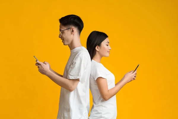 Asian couple standing back to back, using cellphones — Stockfoto