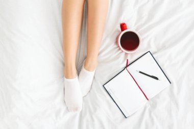 Top view on female legs, notebook and cup of coffee in bed