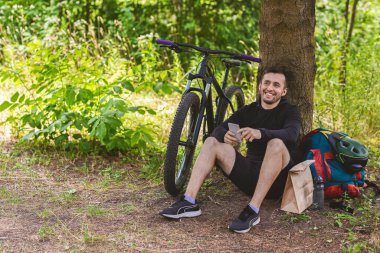 Cheerful cyclist having break under the tree, holding cellphone