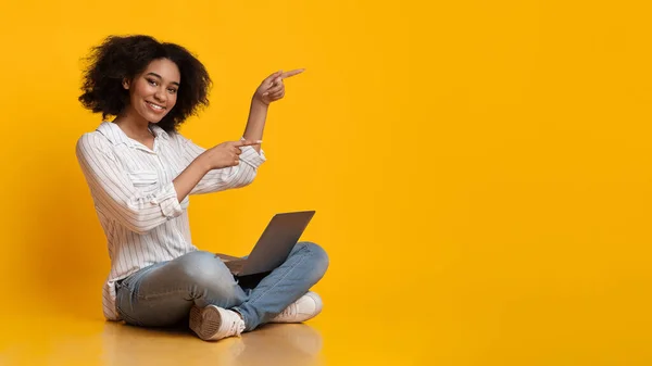 Alegre Afro Girl Sentado en el piso con el ordenador portátil y señalando a un lado —  Fotos de Stock