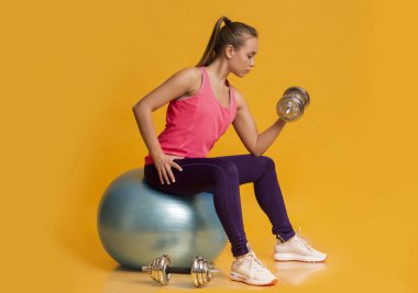 Sporty Woman Exercising With Barbells, Sitting On Fitness Ball In Studio