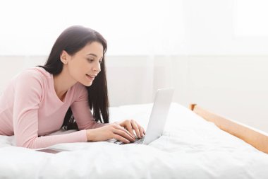 Female journalist typing on laptop, lying on bed at home