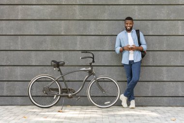 Black Millennial Guy Standing Next To Bike Over Grey Urban Wall