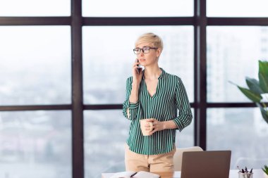 Serious Businesswoman Talking On Cellphone Standing Near Window In Office
