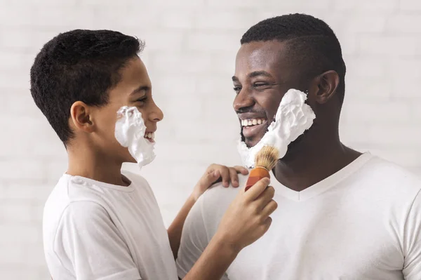 Pequeño hijo negro usando cepillo de afeitar aplicando espuma a la cara de los padres —  Fotos de Stock