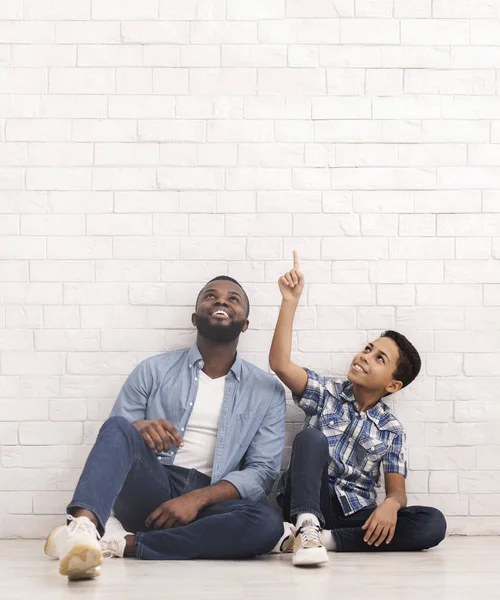 Garoto afro animado sentado no chão com o pai e apontando para cima — Fotografia de Stock