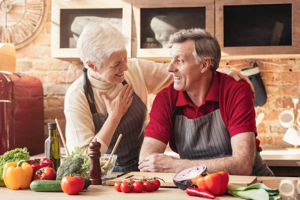 Aged couple laughing and hugging while cooking food in kitchen together — Stok fotoğraf