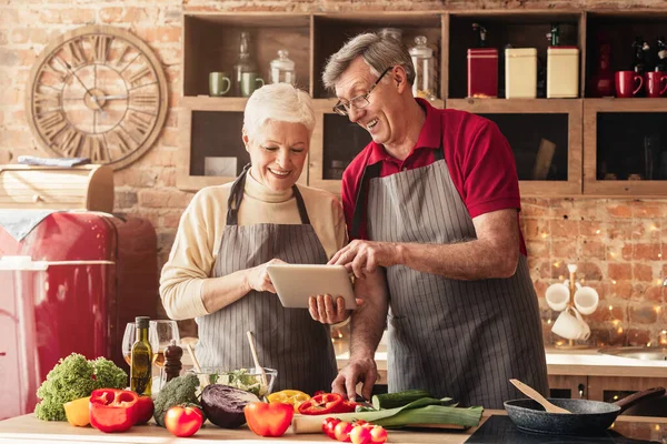 Elderly couple using digital tablet in kitchen, looking for new recipes — Stockfoto