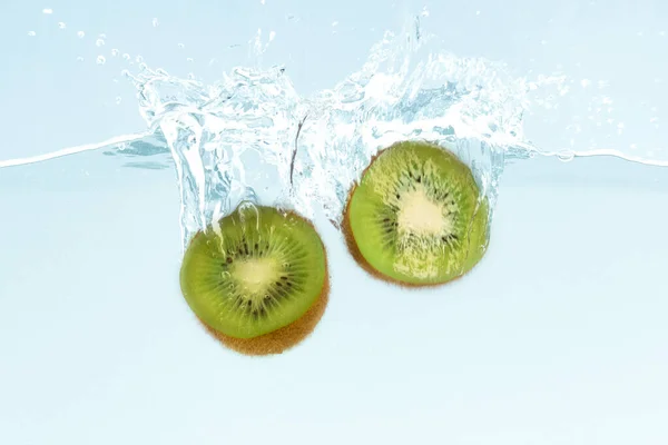 Fresh kiwi halves dropping into clear water with splashes on blue background — Stockfoto