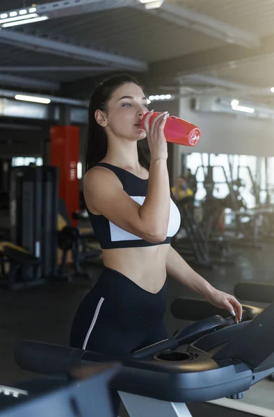 Jovem mulher bebendo água ou batido de proteína enquanto corre em esteira no clube esportivo — Fotografia de Stock