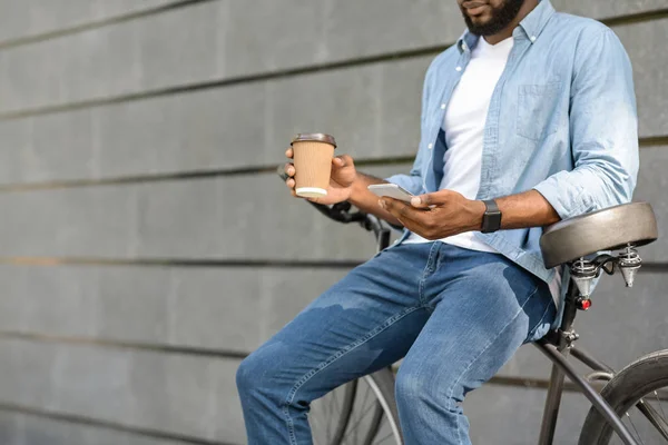 Black millennial guy with smarthone and takeaway coffee sitting on bicycle — 스톡 사진