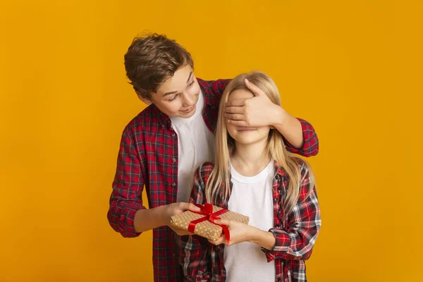 Brother Covering Sisters Eyes And Giving Birthday Gift Over Yellow Background — Stockfoto