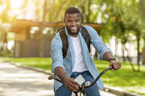 Bonito homem preto com mochila e Smartphone equitação bicicleta, posando ao ar livre — Fotografia de Stock