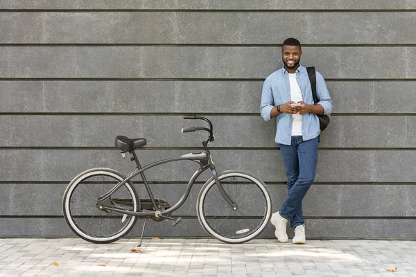 Preto milenar cara de pé ao lado de bicicleta sobre cinza urbano parede — Fotografia de Stock