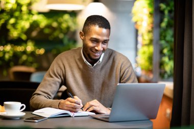 Black business guy taking notes in notebook holding tablet