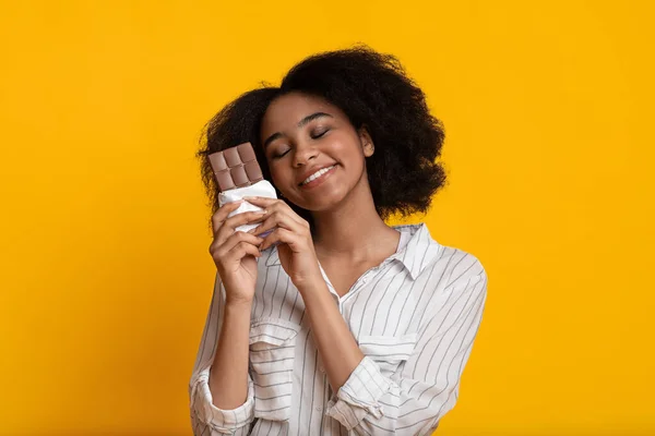 Happy african american woman hugging chocolate bar, standing over yellow background — Stockfoto