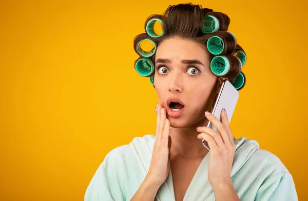 Scioccato ragazza con bigodini parlando sul cellulare in piedi, sfondo giallo — Foto Stock