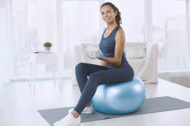 İnce Latin kadın, evde dambıllarla ve egzersiz topuyla çalışıyor, boş alan.