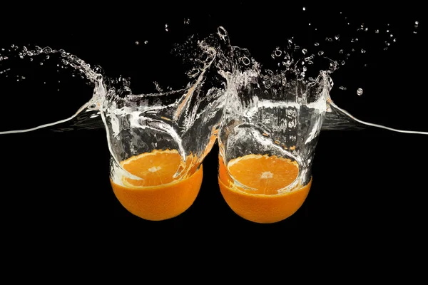 Mitades de naranja dulce cayendo en agua transparente con salpicadura sobre fondo negro — Foto de Stock