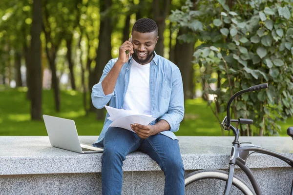 Homem negro sentado no parque conversando no celular e verificando documentos — Fotografia de Stock