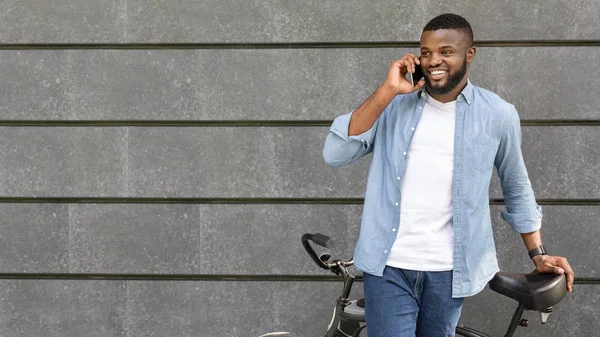 Радісний чорний хлопець стоїть з велосипедом і розмовляє на смартфоні на відкритому повітрі — стокове фото