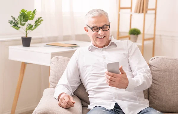 Cheerful aged man using smartphone sitting on sofa — Stock Photo, Image