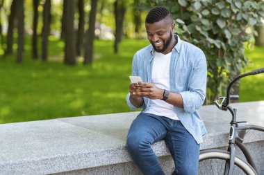 Neşe dolu siyah adam bisikletini bankın yanına park etmiş ve parkta akıllı telefon kullanıyor.