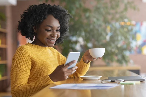 Feliz afro mulher bebendo café e usando telefone celular — Fotografia de Stock