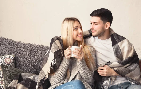 Sonriente pareja joven con bebidas calientes abrazándose bajo una manta caliente en casa, espacio vacío — Foto de Stock