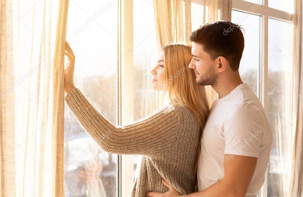 Beautiful millennial couple standing near window at home