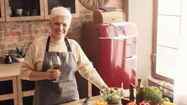 Pozytywny starszy kobieta z kieliszkiem wina w rękach pozowanie w kuchni — Zdjęcie stockowe