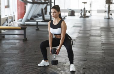 Dambılı genç bir kadın spor salonunda çömeliyor.