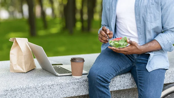 Unrecognizable Black Man Eating Salad, Drinking Coffee And Using Laptop Outdoors