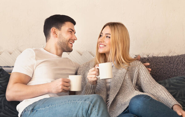 Happy couple with hot drinks relaxing on sofa at home