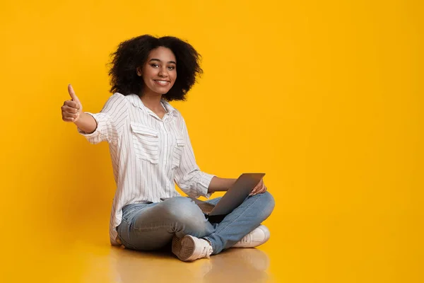 Посміхаючись Афро дівчина сидить з ноутбуком і показує великий палець вгору жест — стокове фото