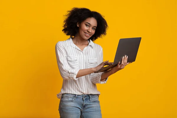 Online Μαθήματα. Χαμογελώντας μαύρη γυναίκα χρησιμοποιώντας φορητό υπολογιστή, στέκεται σε κίτρινο φόντο — Φωτογραφία Αρχείου