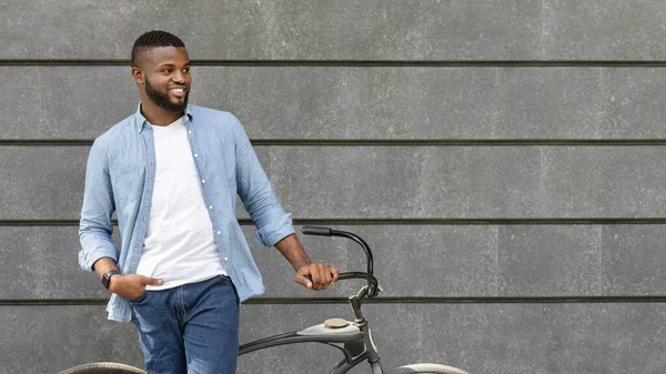 Bonito homem negro de pé com bicicleta contra a parede urbana, olhando para longe — Fotografia de Stock