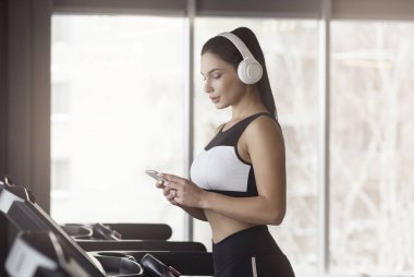 Charming girl with smartphone and headphones listening to music on treadmill, copy space
