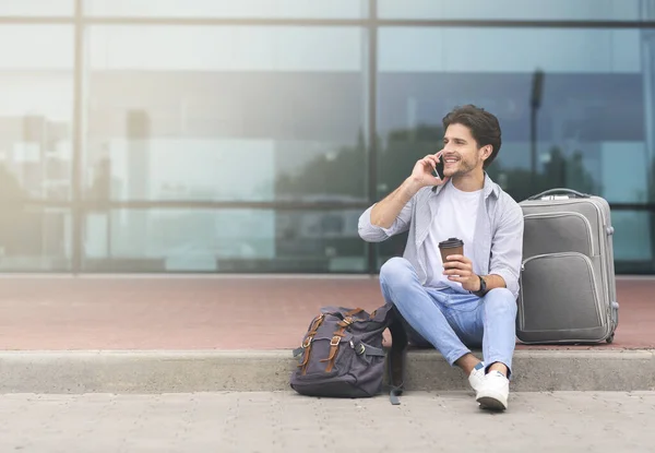 Homem alegre sentado com bagagem perto do aeroporto e conversando no celular — Fotografia de Stock