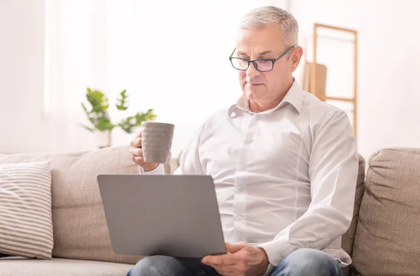 Anciano mirando a la computadora portátil y tomando café — Foto de Stock