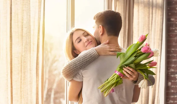 Mujer sentimental con flores abrazando a su novio amoroso cerca de la ventana en casa, panorama. Espacio para texto — Foto de Stock