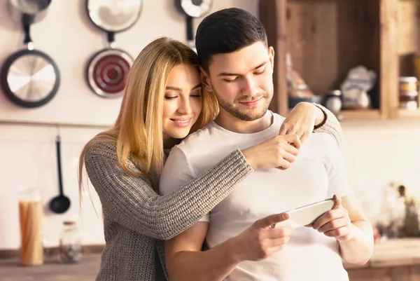 Romance todos os dias. Menina loira e seu namorado bonito com abraço smartphone na cozinha — Fotografia de Stock