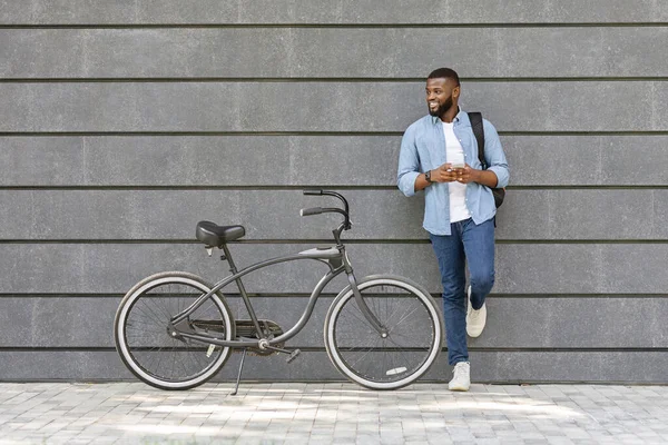 Chico afro guapo con teléfono inteligente de pie cerca de la bicicleta, apoyado en la pared — Foto de Stock