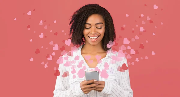 African American woman browsign through social media on mobile phone with hearts flying around, pink background — стокове фото