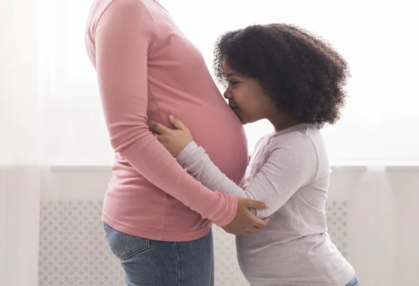 Adorable little afro daughter kissing mothers pregnant belly at home — Zdjęcie stockowe