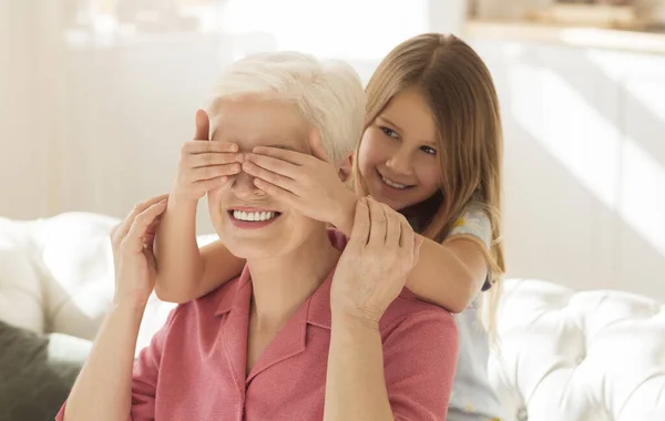 Adorable girl closing her grannys eyes at home, playing GUESS WHO game — Stock Photo, Image