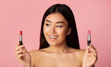 Asian Girl Holding Two Lipsticks Choosing Standing On Pink Background
